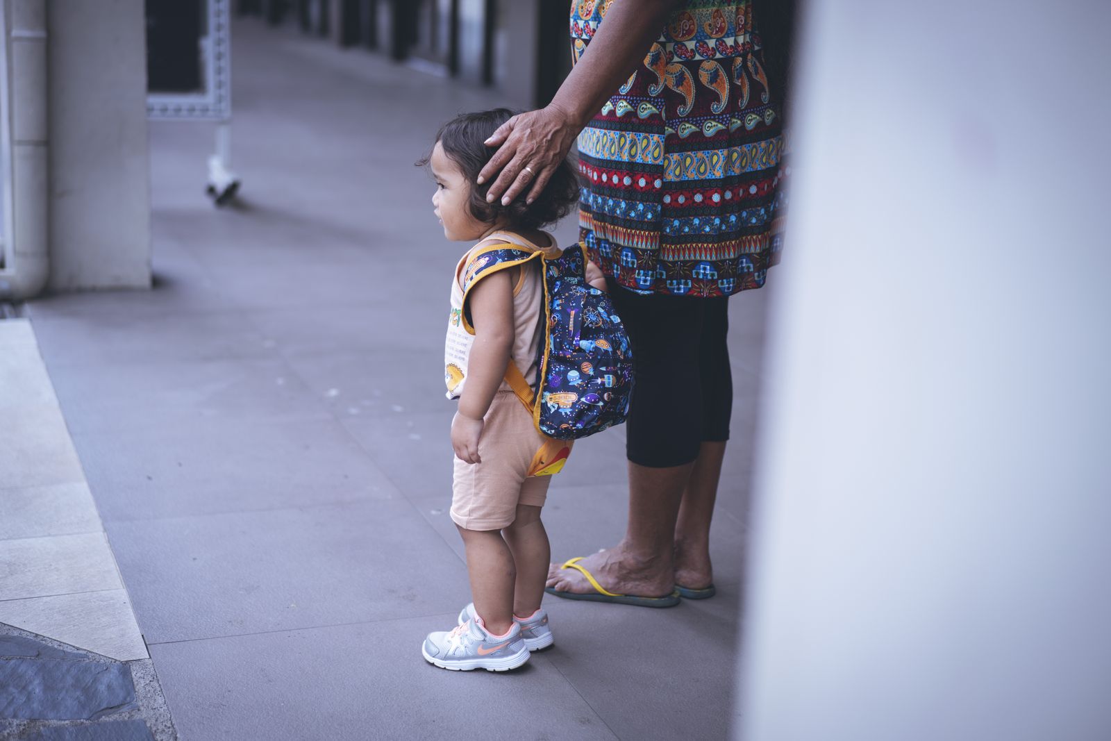 Toddler with a colorful backpack standing beside a caregiver at daycare drop-off