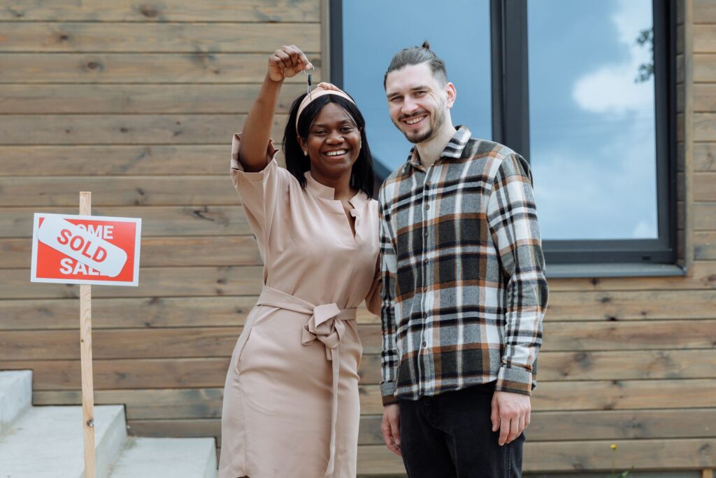 Smiling couple holding up the keys in front of their new house with a sold sign